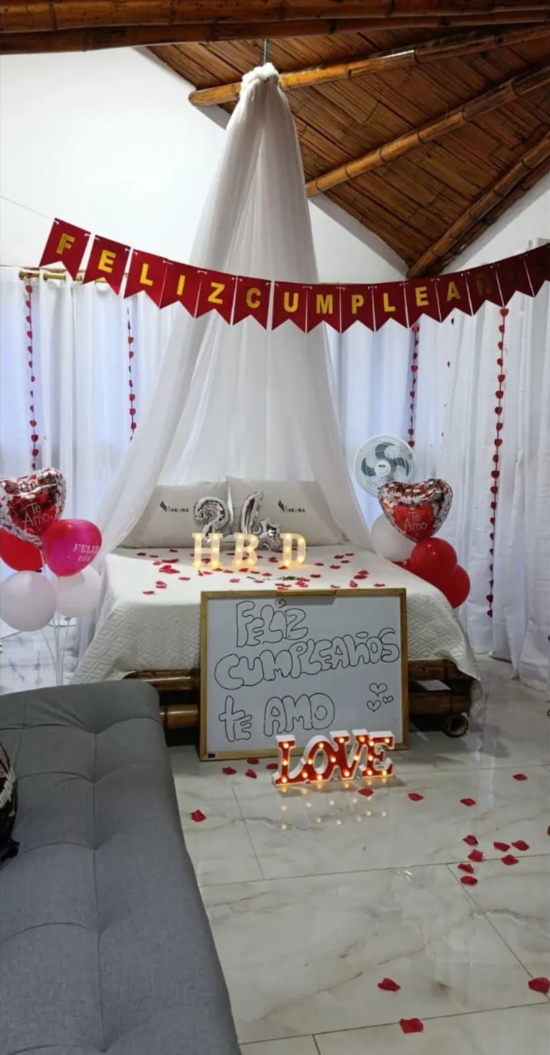 Cama con toldo blanco decorada para un cumpleaños, con globos, pétalos de rosa y letras de luz